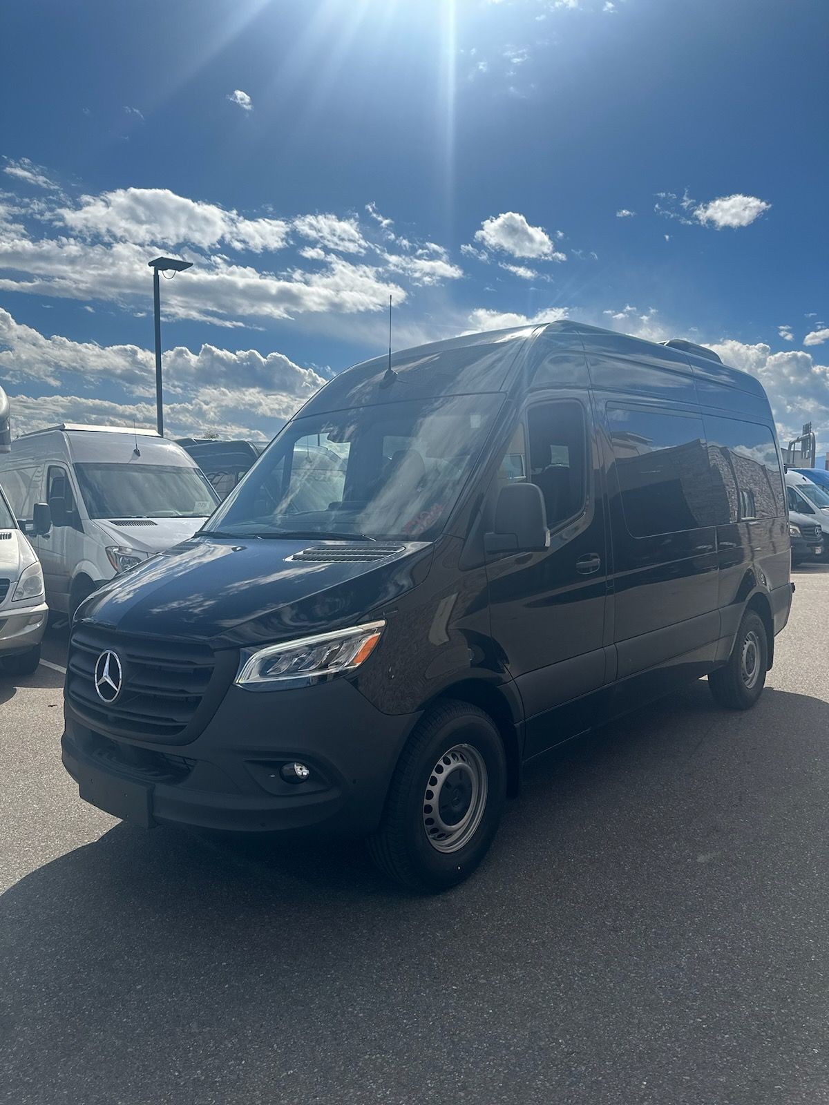 Black van parked outdoors under a clear blue sky with white clouds and sunlight shining down.